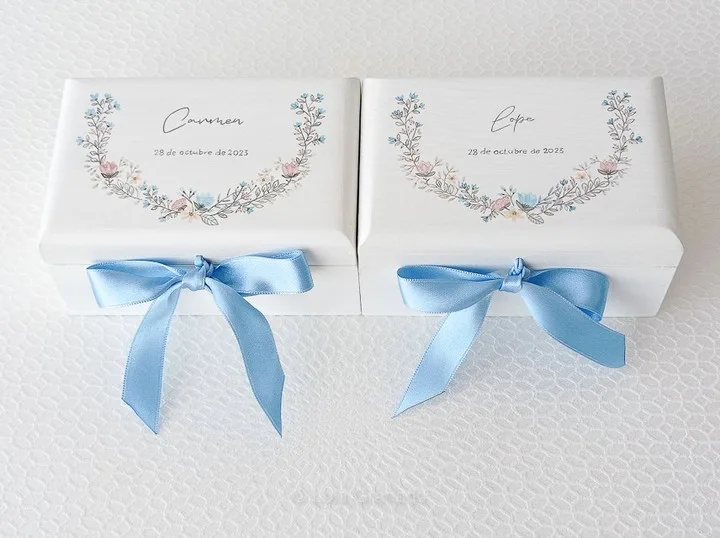 Dos cajas blancas en forma de arca, con flores en tonos pastel y lazos celestes